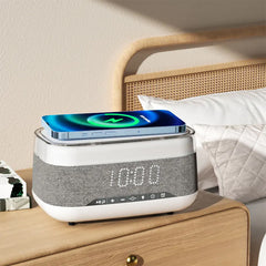 Image of an alarm clock with a phone charging on top of it on a nightstand next to a bed. The alarm clock is black and has a digital display. The phone is white and is sitting in a wireless charging dock. The nightstand is made of wood and has a lamp on it. The bed is made of white sheets and has a blue blanket on it.